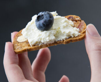 a photo os a hand holding melba toast topped with crowdie and a blueberry